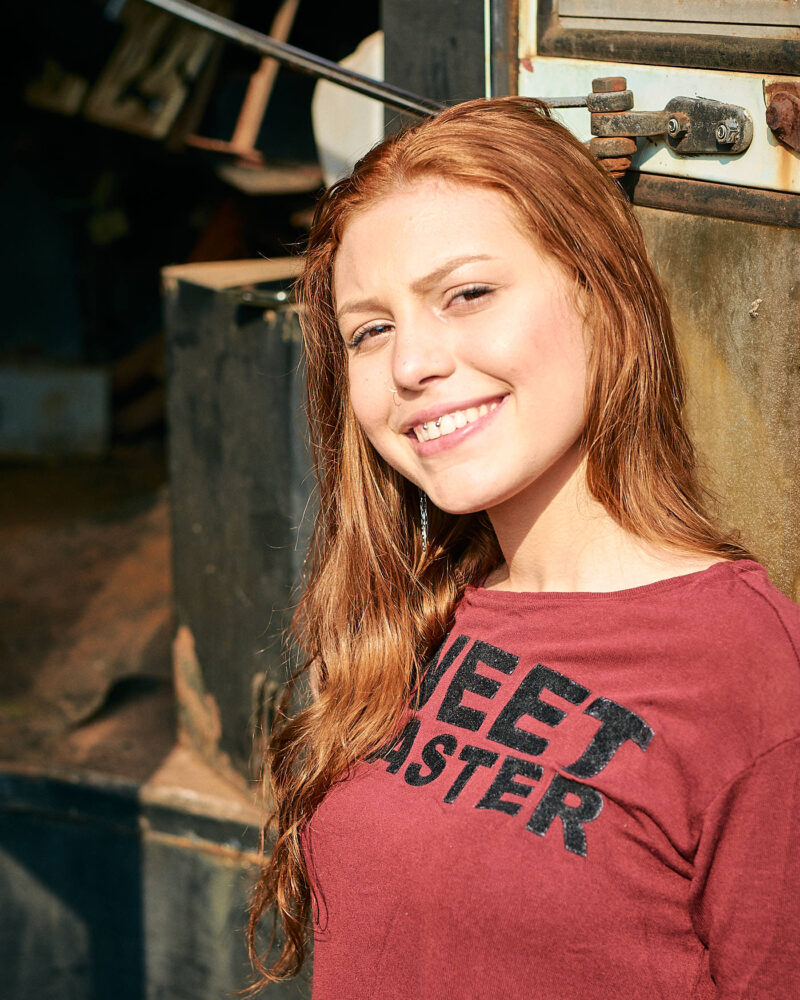 Mulher ruiva sorrindo sob o sol da tarde, encostada na porta de um ônibus velho