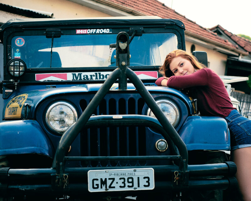 Mulher ruiva posando apoiada em um jipe azul, em um ensaio externo