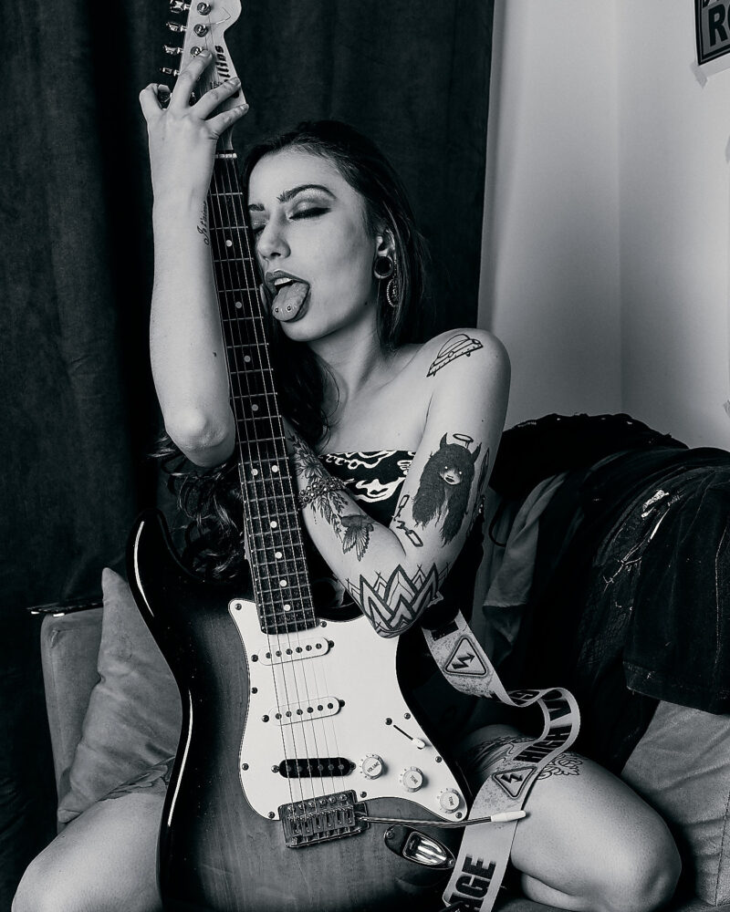 Mulher posando com uma guitarra e a língua de fora, em cima do sofá