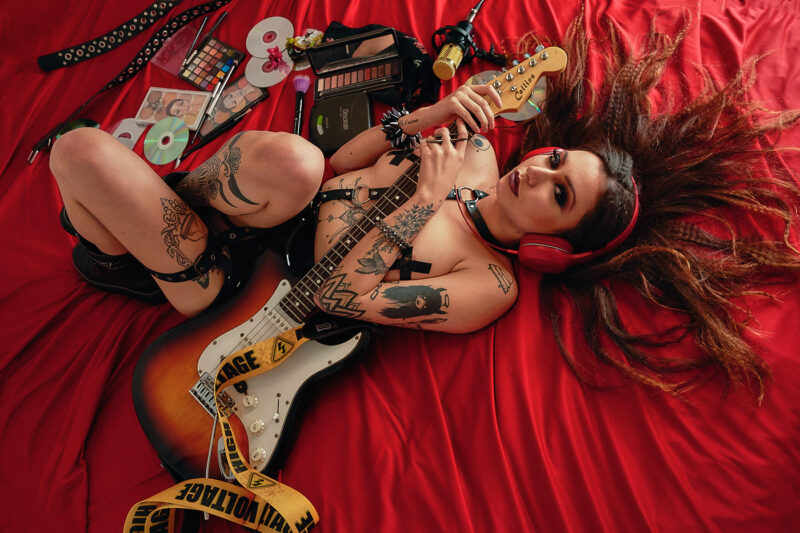 Mulher deitada sobre lençol vermelho, sem blusa, segurando uma guitarra, em um ensaio sensual